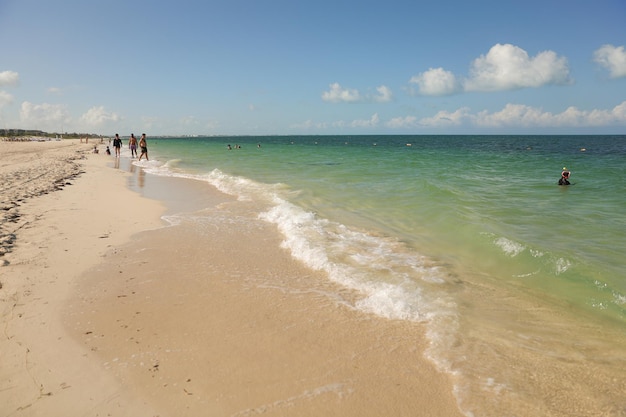 La playa está llena de gente caminando sobre la arena y el agua es azul y verde.