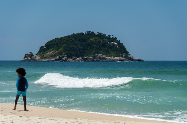 Playa de Grumari cerca de Barra da Tijuca en Río de Janeiro, Brasil. Día soleado con cielo azul, agua clara. Colinas y naturaleza alrededor. Niño negro, fuera de foco, jugando en la arena.