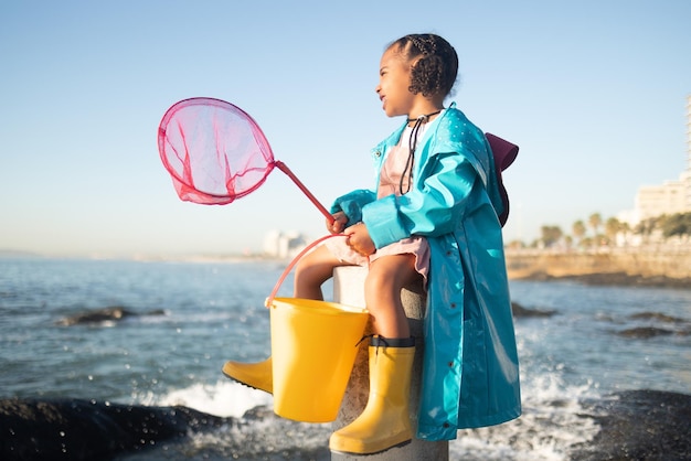 Playa de niñas y red de pesca con agua de cubo y piscina de rocas para aventuras al aire libre con botas, sonrisa y océano Mar de niños negros y feliz por las olas de peces o el mar en la naturaleza, el aprendizaje y el sol