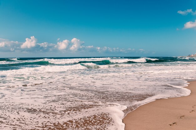 Foto playa del océano con huellas en la arena amarilla, cielo azul, nubes blancas, olas turquesas