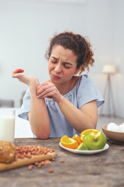 Plötzlicher Ausschlag. Heranwachsende Frau, die an einem Esstisch sitzt und besorgt und genervt aussieht und erste Symptome eines Hautausschlags erfährt, der durch eine Nahrungsmittelallergie verursacht wird