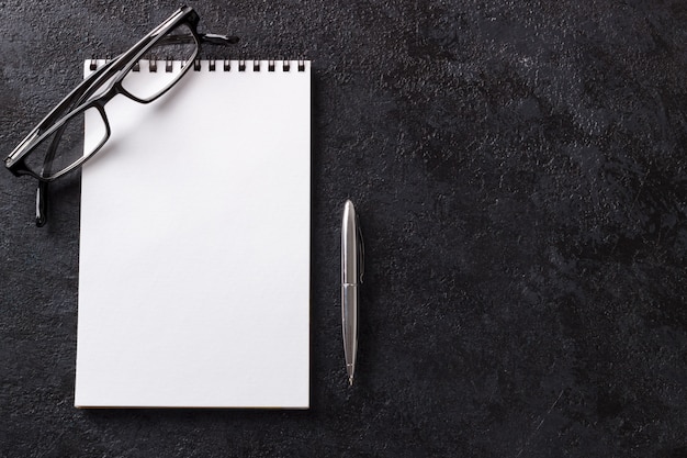 La pluma y los vidrios en blanco de la libreta en la tabla moderna de madera oscura imitan encima de visión superior