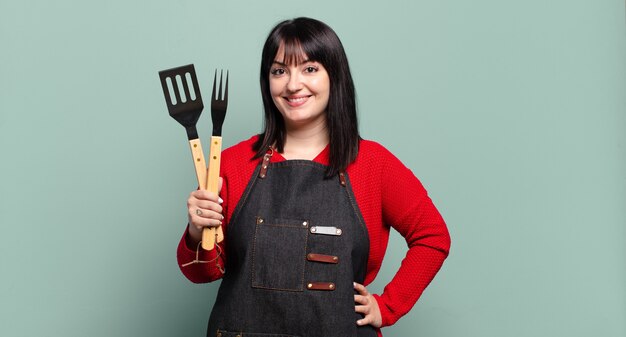 Plus Größe hübsche Frau, die glücklich mit einer Hand auf Hüfte und selbstbewusster, positiver, stolzer und freundlicher Haltung lächelt