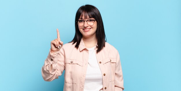 Plus Größe hübsche Frau lächelnd und freundlich aussehend, zeigt Nummer eins oder zuerst mit der Hand nach vorne, Countdown