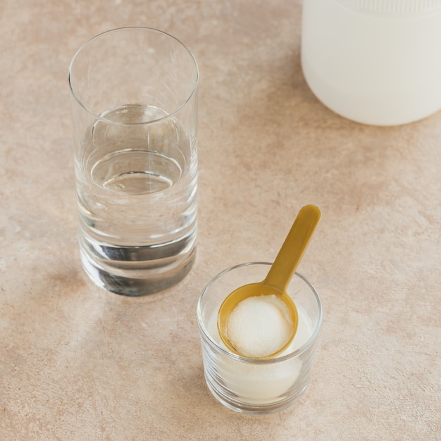 Pó de colágeno em uma tigela, um copo de água e uma colher de medida em um fundo bege claro. ingestão extra de proteína. conceito de suplemento de beleza e saúde natural.