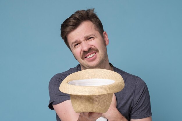 Pobre joven sosteniendo sombrero y pidiendo dinero sobre fondo azul.