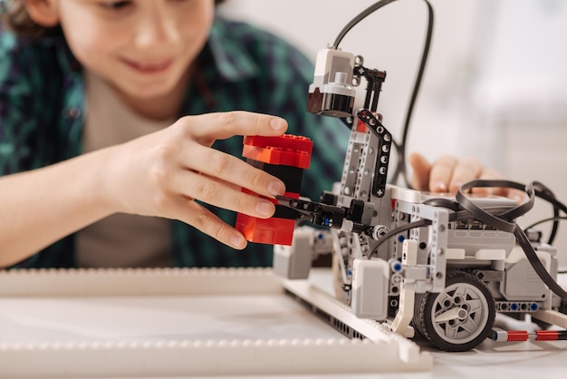 Época da invenção da tecnologia. Menino habilidoso e animado e sorridente sentado na sala de aula enquanto estuda e constrói um robô