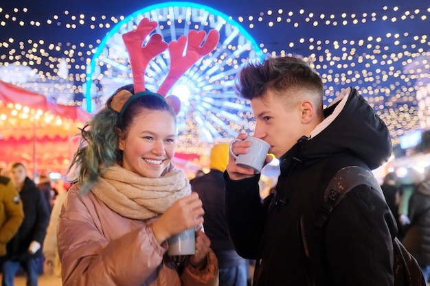 Época de Natal, feriados de ano novo. Jovens, dois adolescentes se divertindo no mercado de Natal, bebendo chá quente em canecas, conversando, rindo, guirlandas de fundo da cidade à noite, roda gigante