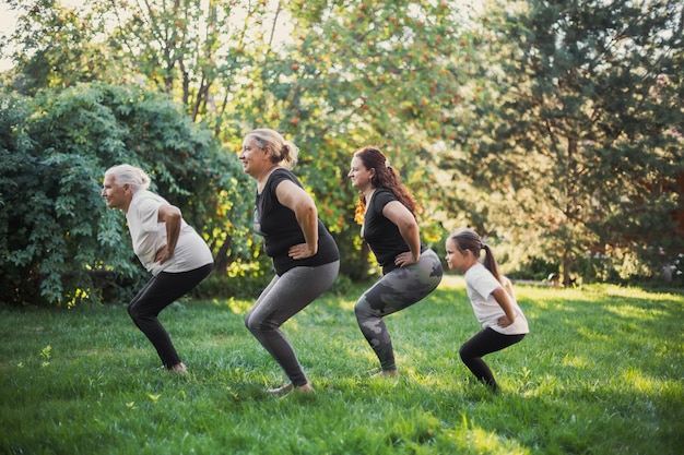 Pocas generaciones de mujeres de una familia en cuclillas haciendo ejercicios físicos de la misma manera juntas en el patio trasero en un prado lleno de hierba verde y árboles Pasando tiempo juntos