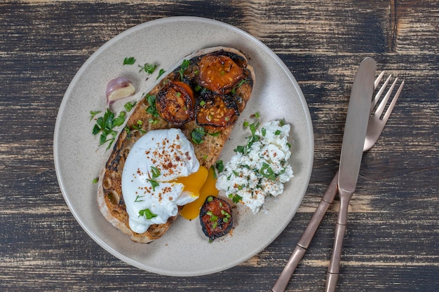 Pochiertes Ei auf einem Stück Brot mit gebratenen roten Tomaten, Knoblauch und Hüttenkäse auf einem Teller aus nächster Nähe