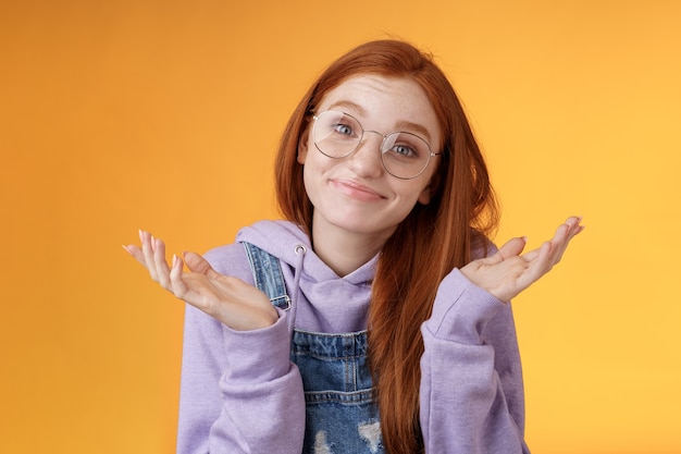 Poco. Atractiva y tonta pelirroja despreocupada insegura y despreocupada linda mujer con gafas geek sonriendo con satisfacción incierta encogiéndose de hombros con las manos extendidas hacia los lados sin idea de qué hacer en el futuro, fondo naranja.