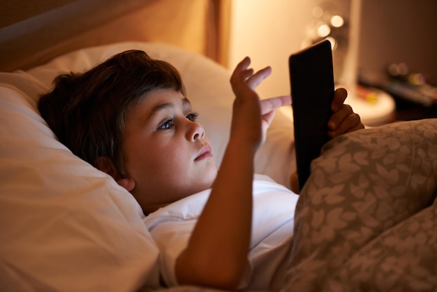 Un poco de lectura ligera antes de acostarse Foto de un niño jugando en una tableta mientras está acostado en la cama