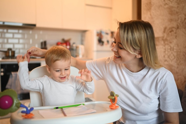 Poco lindo bebé niño rubio sentado en una silla de bebé aprendiendo a dibujar hermosa joven mamá y