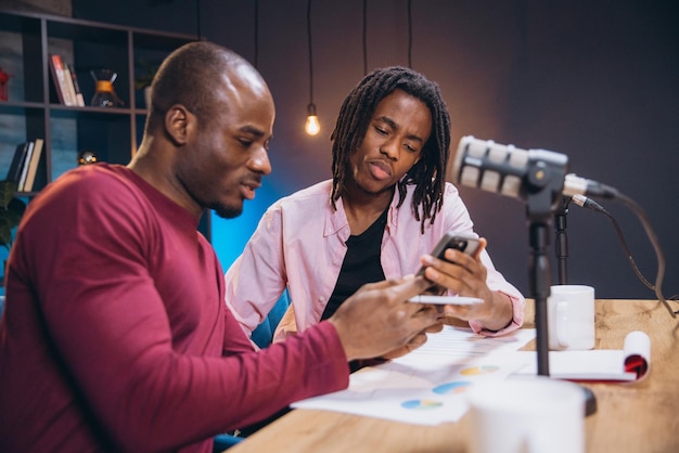 Podcasters gravando episódio referenciando smartphone na mesa com microfone e papéis nas proximidades