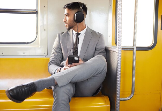 Podcasts ajudam a passar o tempo. Foto de um jovem empresário usando fones de ouvido e usando um celular enquanto olhava pela janela em um trem durante seu trajeto.