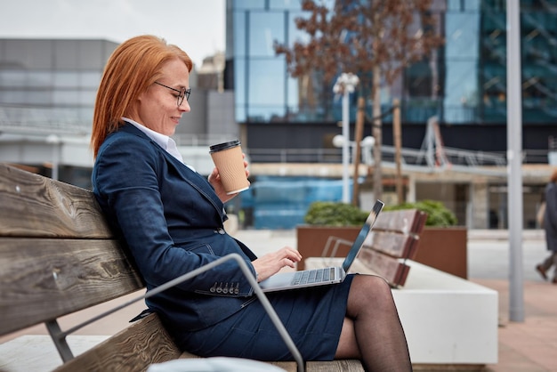 Poderosa mujer de negocios que se hace cargo afuera con su computadora portátil tomando un café para llevar