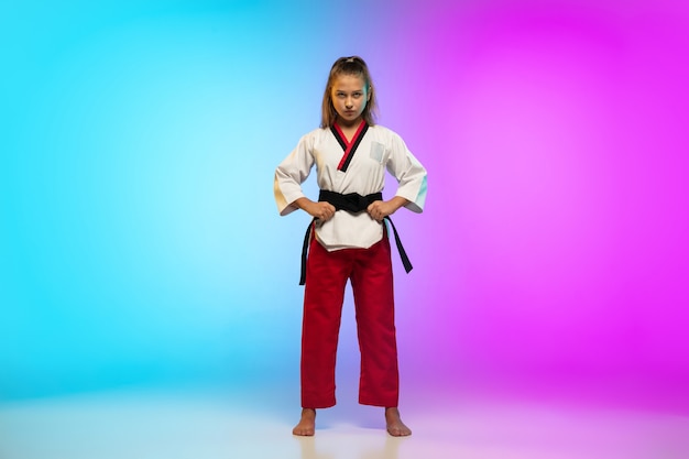 Poderoso. karatê, garota de taekwondo com faixa preta isolada em fundo gradiente em luz de néon. pequeno modelo caucasiano, garoto do esporte, treinamento em movimento e ação. esporte, movimento, conceito de infância.