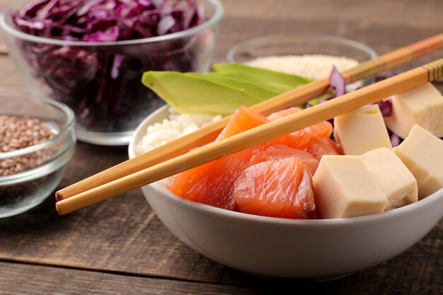 Poke bowl comida havaiana. um prato com arroz, salmão, abacate, repolho e queijo, perto de gergelim. em uma mesa de madeira marrom.