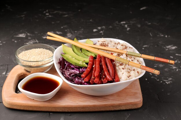 Poke bowl comida hawaiana. un plato de arroz, salmón, aguacate, repollo y queso. cerca de la mofeta y la salsa de soja sobre un fondo negro.