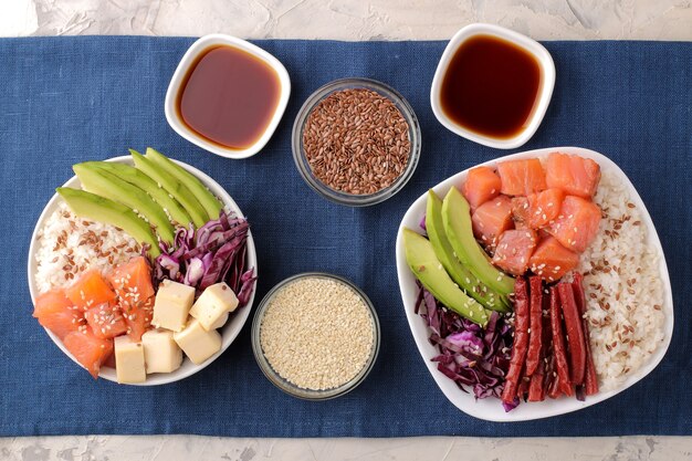 Foto poke bowl comida hawaiana. un plato con arroz, salmón, aguacate, repollo y queso junto a un zorrillo y salsa de soja sobre una servilleta azul sobre un fondo claro. vista superior