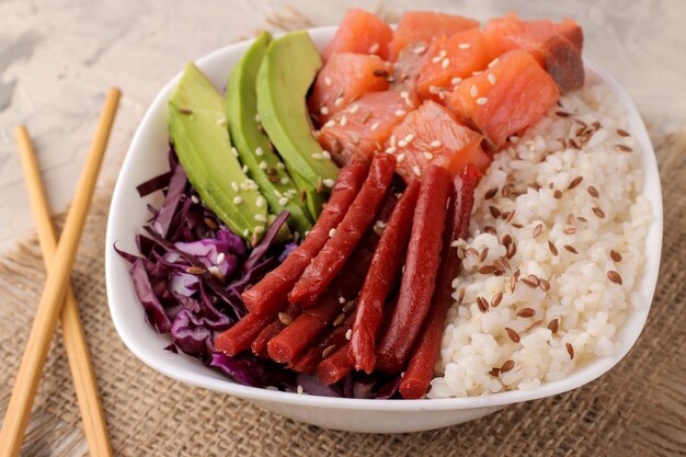 Poke bowl comida hawaiana. un plato con arroz, salmón, aguacate, repollo y queso sobre un fondo claro con palillos.