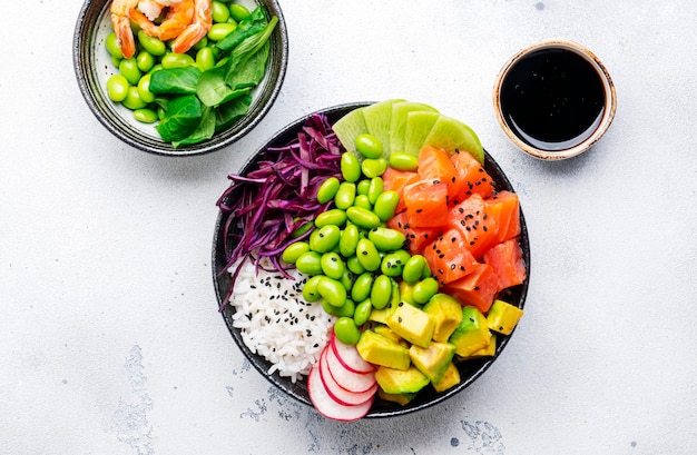 Poke bowl hawaiano con salmón, aguacate, rábano, frijoles edamame, repollo rojo y arroz blanco, salsa de soja, lima y aderezo de sésamo, vista superior del fondo de la mesa blanca