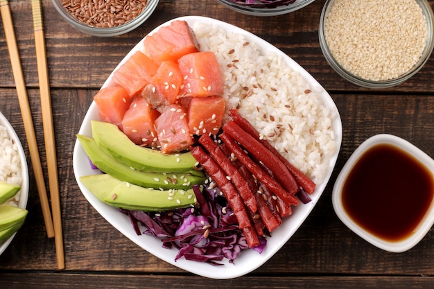 Poke Bowl hawaiianisches Essen. ein Teller mit Reis, Lachs, Avocado, Kohl und Käse, dazu Sesam und Sojasauce. auf einem braunen Holztisch. Ansicht von oben