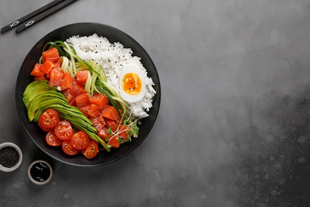 Poke bowl con salmón, aguacate, huevo y arroz sobre fondo oscuro con palillos. Vista superior