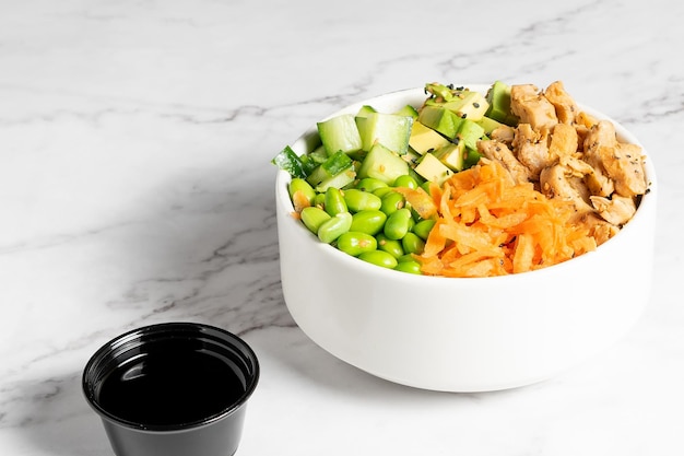 Poke de pollo, zanahoria, pepino y frijoles servido en un bol blanco sobre una mesa de mármol.