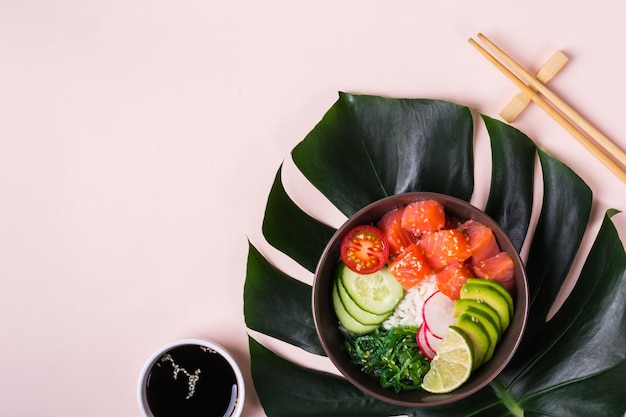 Poke de salmón hawaiano con aguacate sobre fondo rosa con hojas tropicales.