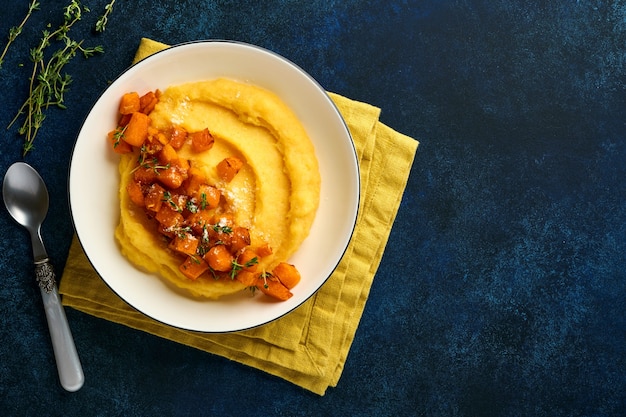 Polenta com manteiga, abóbora, alho, queijo parmesão e salgado em uma tigela branca sobre fundo de concreto azul escuro. Comida italiana básica tradicional, comida vegana. Vista do topo. Copie o espaço.