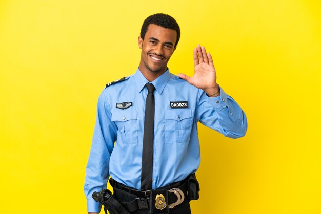 Policial afro-americano sobre fundo amarelo isolado saudando com a mão com expressão feliz