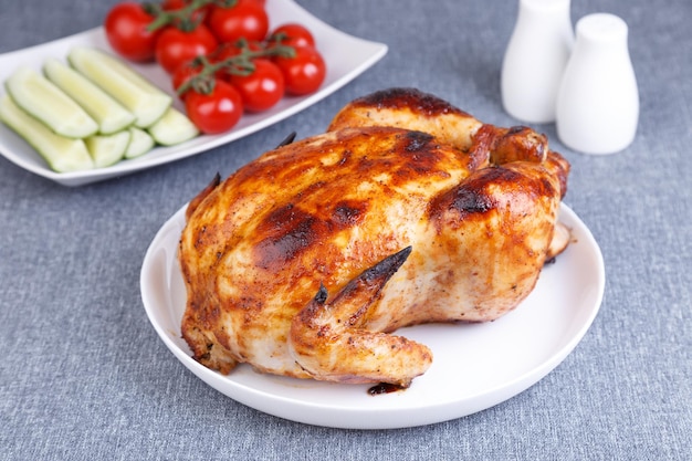 Pollo al horno entero con una corteza deliciosa Un plato tradicional En el fondo hay pepinos y tomates cherry en un plato blanco Primer plano enfoque selectivo fondo gris