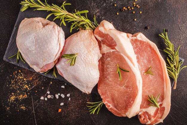 Pollo y cerdo crudos en pizarra con las especias. Fondo oscuro, vista desde arriba, copia espacio.