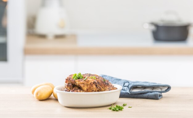 Pollo entero asado en plato blanco con patatas crudas en mesa cocinada. Interior de una casa en el fondo.