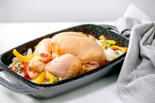 Foto pollo entero crudo orgánico crudo con verduras en rodajas pimiento, cebolla y hierbas en bandeja de horno con servilleta textil gris, listo para cocinar. mesa de mármol blanco.