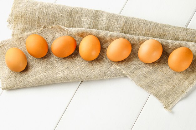 El pollo marrón crudo eggs en una fila en la arpillera en una tabla de madera blanca. Ingredientes para cocinar.