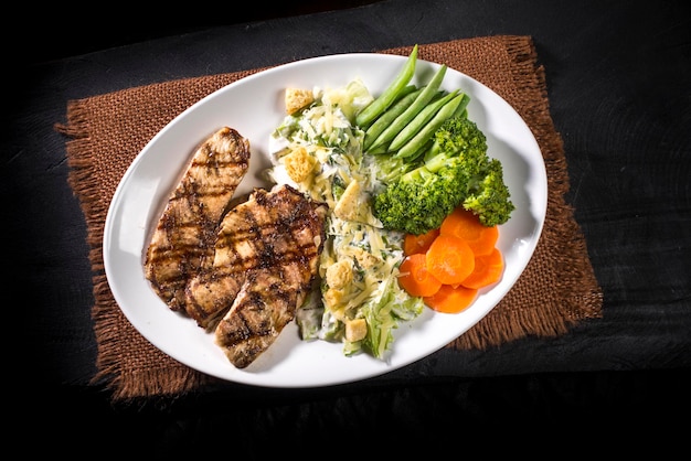 Pollo a la parrilla, ensalada césar y verduras sobre tela marrón y vista superior de madera negra