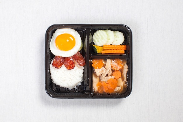 Foto pollo con salsa con huevo frito sobre arroz puesto en una caja de plástico negra, poner sobre un mantel blanco, caja de comida, comida tailandesa.