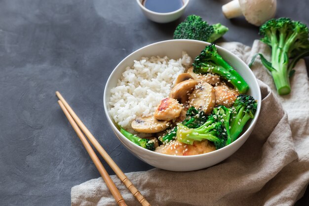 Pollo teriyaki, brócoli y champiñones salteados con arroz blanco en un recipiente sobre hormigón gris.