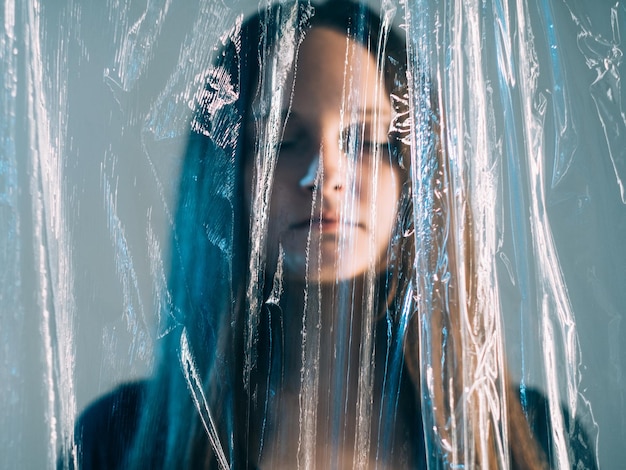 Poluição plástica. Reciclagem de lixo. Problema de ecologia. Salve o planeta. Retrato de borrão frio mulher sem emoção com olhos fechados se afogando sob filme de polietileno transparente isolado sobre fundo azul.