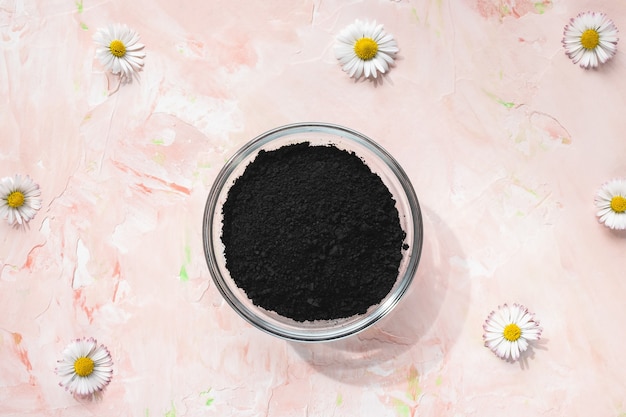 Polvo de carbón activado negro en un recipiente de vidrio sobre fondo floral rosa pastel. Ingrediente para belleza, cosmética, tratamiento de spa, concepto de desintoxicación. Vista superior, espacio de copia