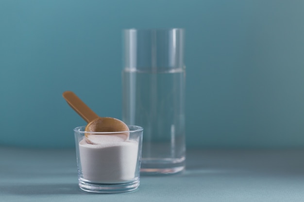 Polvo de colágeno en un recipiente, un vaso de agua y una cuchara medidora en azul claro