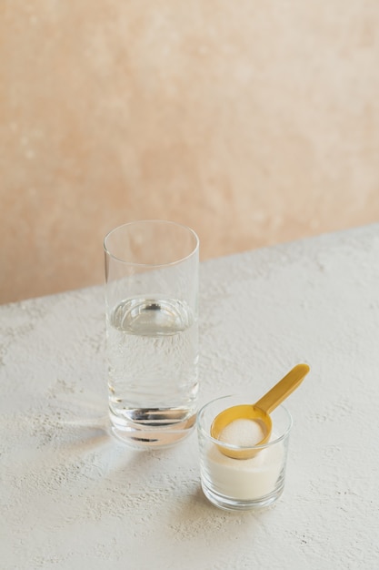 Polvo de colágeno en un recipiente, un vaso de agua y una cuchara medidora en blanco
