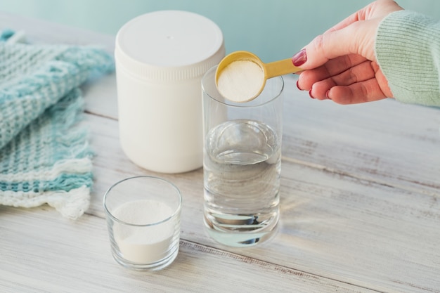 Polvo de colágeno en un recipiente, un vaso de agua y una cuchara medidora en una madera blanca.