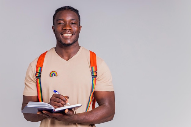 Polyethnischer Mann in beigem T-Shirt mit orangefarbenem Rucksack, der Buch und Notizbücher im grauen Hintergrundstudio hält