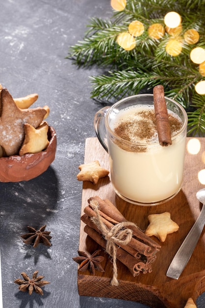 Ponche de huevo de bebida tradicional de Navidad en vaso de vidrio, galletas con forma de corazón sobre tablero de cocina de madera y ramas de abeto con luces bokeh sobre fondo gris. Acogedora casa navideña. Orientación vertical.