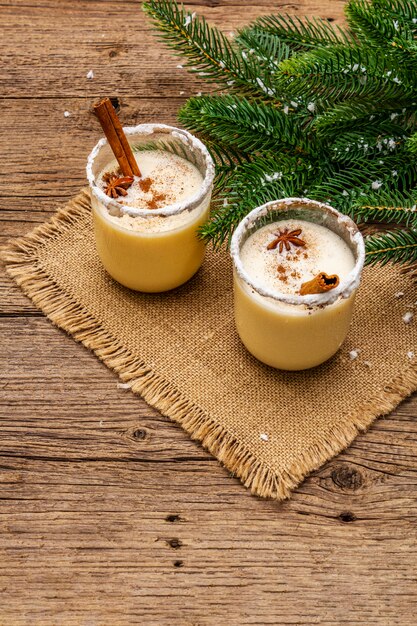 Ponche de huevo cóctel en dos vasos dispuestos con decoración de Navidad en la mesa de madera vieja Evergreen fer rama de árbol, nieve artificial, servilleta de tela de saco