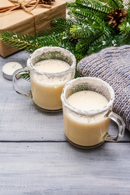 Ponche de huevo cóctel en dos vasos taza dispuesta con decoración de navidad en la mesa de madera clara Rama de abeto perenne, regalo, jersey acogedor, nieve artificial