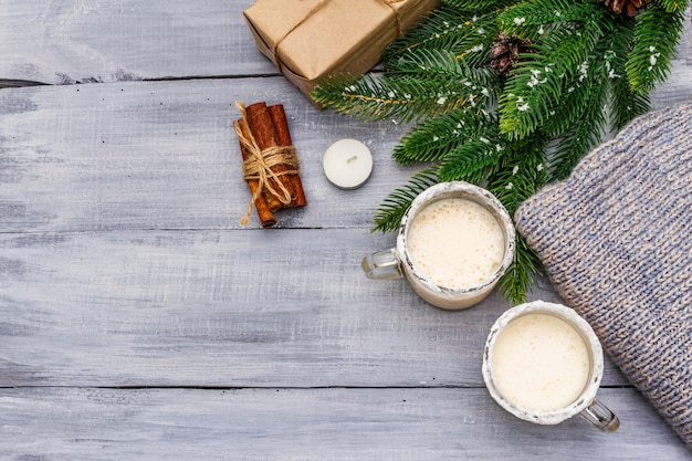 Ponche de huevo cóctel en dos vasos taza dispuesta con decoración de navidad en la mesa de madera clara Rama de abeto perenne, regalo, jersey acogedor, nieve artificial